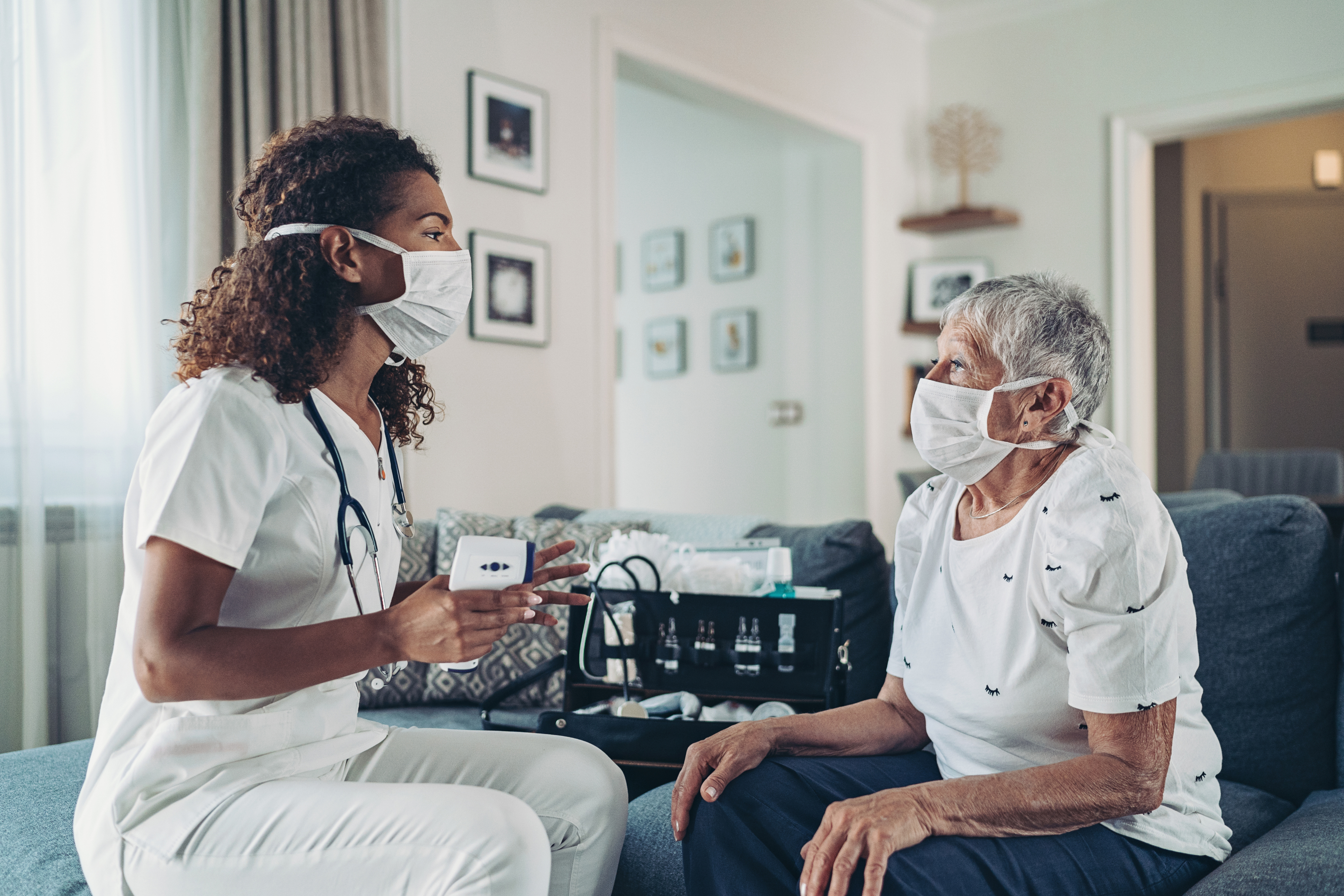 Health practitioner and elderly woman chatting on couch with mask. 