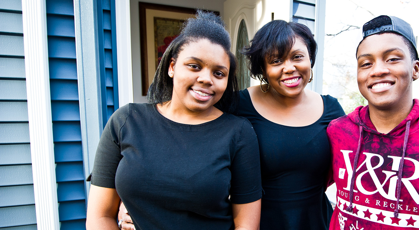 Two Black women and man, smiling, standing in front of a house