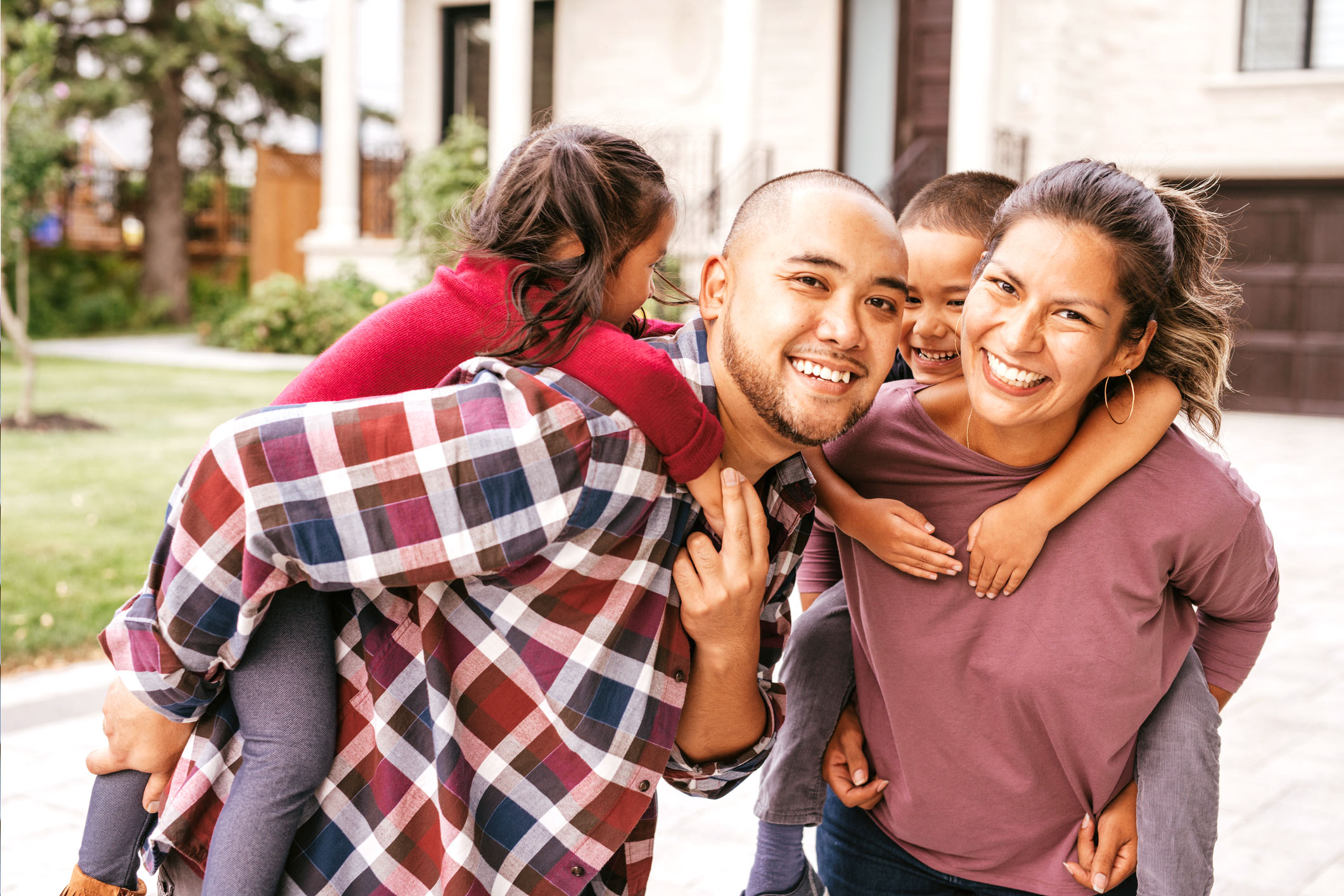 Mother and father smiling with their children climbing on their backs in front of their home