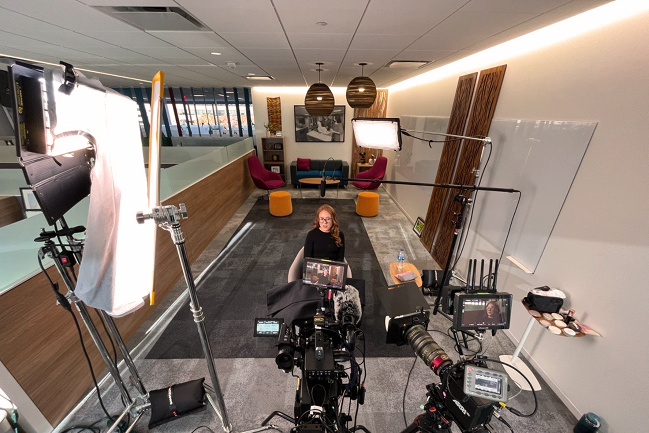 Aerial perspective photo showing multiple cameras and lighting focused on a person sitting in a chair with casual seating and artwork behind them.