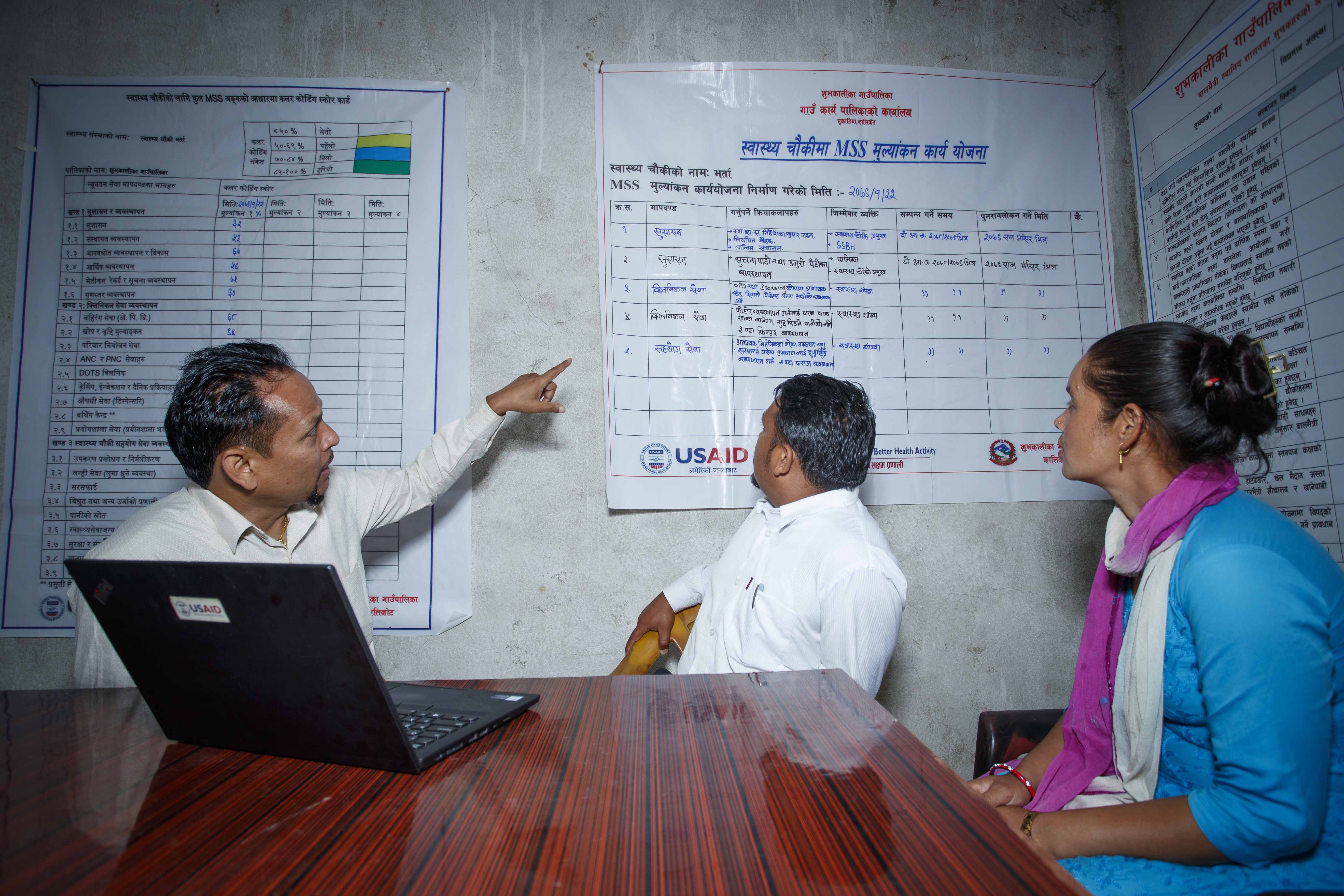 Three people referring to a chart on a wall in Nepal