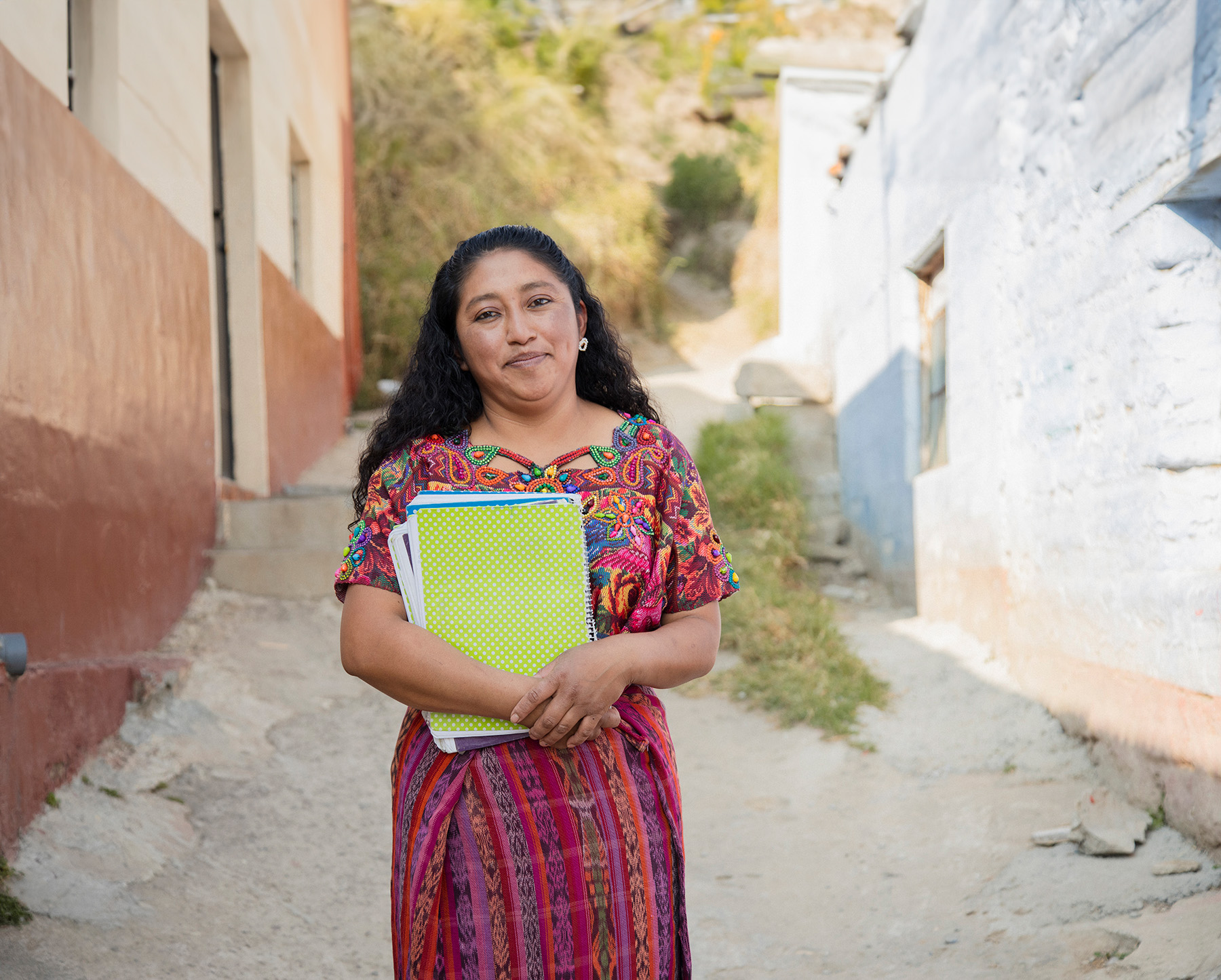 A woman holding notebooks as she stand in the street between buildings.