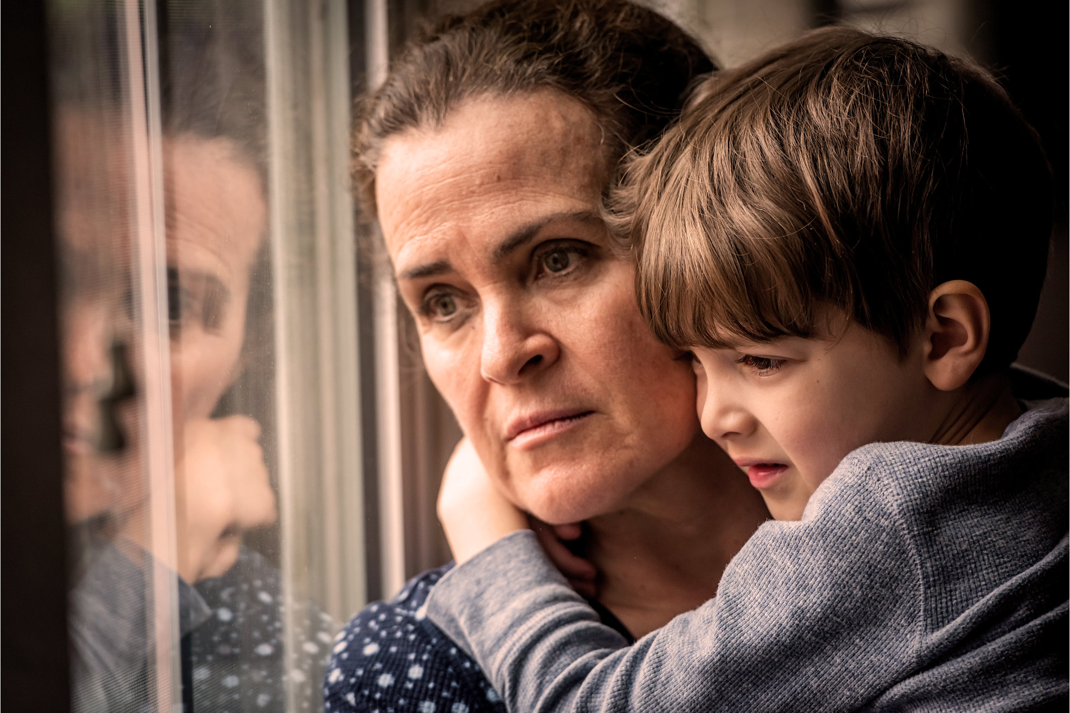 mother looking forlorn looking out a window with her young son in her arms