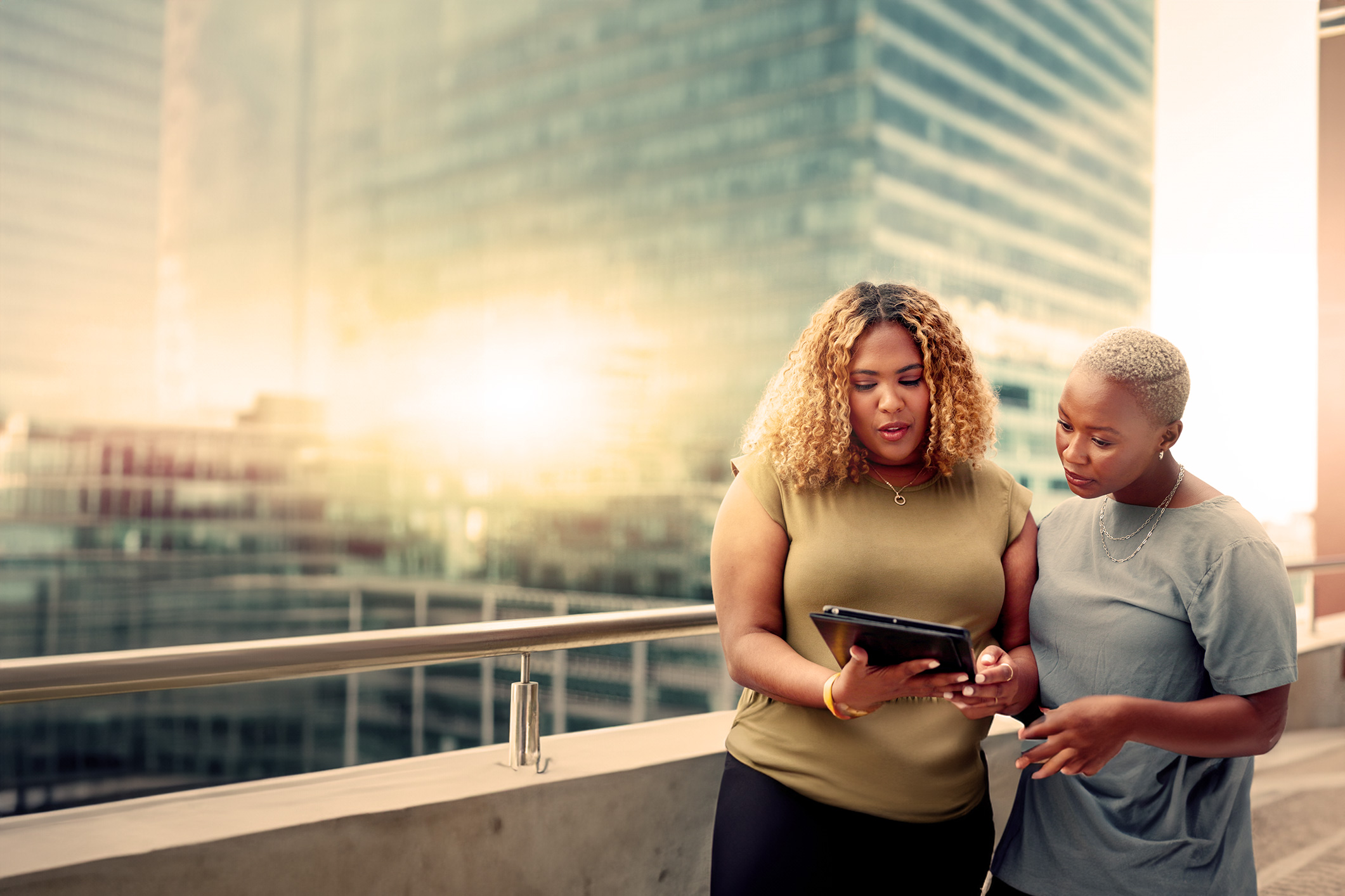 Two POC women standing on an office balcony discussing information on a tablet