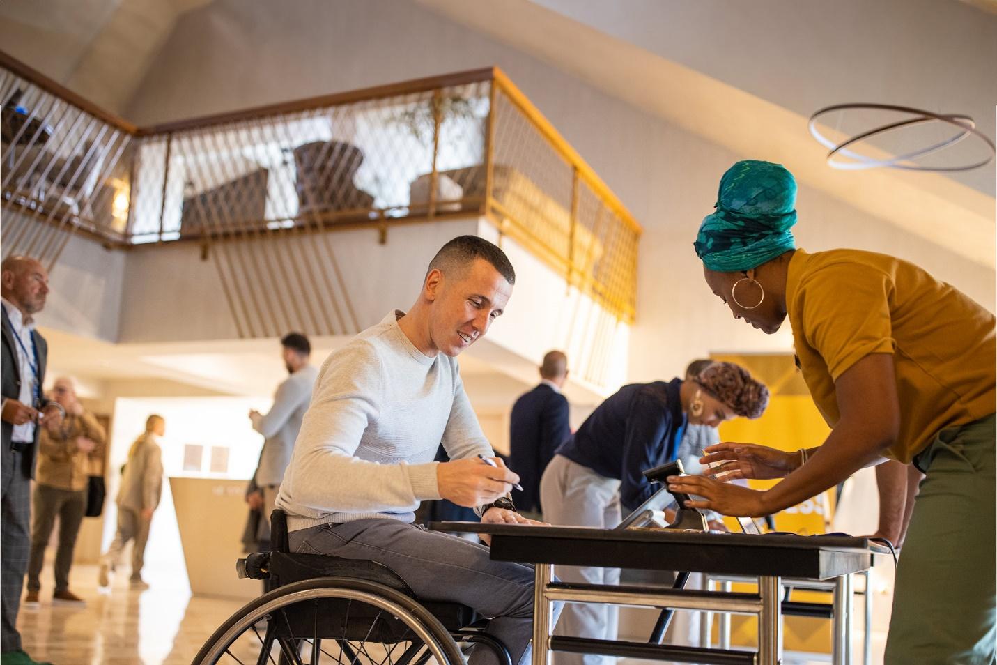 A person in a wheelchair signing in at a conference registration table while conference specialist prepares his badge and conference program