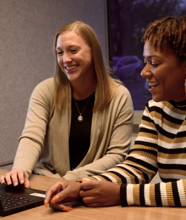 Two women, both smiling, sit and convere while one uses a laptop at the office.