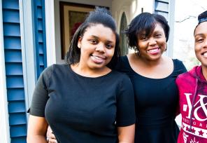 Two Black women and man, smiling, standing in front of a house