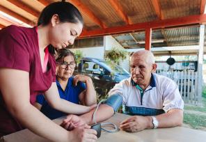 Rural medical visitor woman checking blood pressure of a senior latin male farmer patient, with a monitor