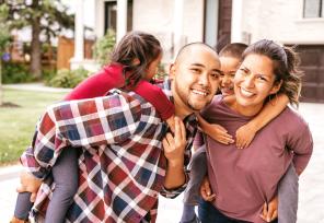 Mother and father smiling with their children climbing on their backs in front of their home