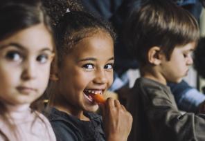 young girl sitting with other kids, smiling and eating a carrot