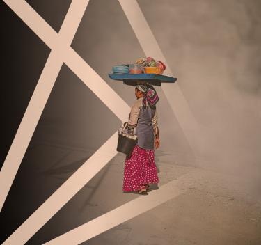 African woman walking with a bucket under her arm of supplies while balancing a tray with bowls of grain and vegetables on her head.