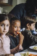young girl sitting with other kids, smiling and eating a carrot