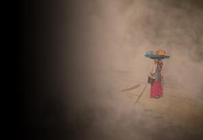 African woman walking with a bucket under her arm of supplies while balancing a tray with bowls of grain and vegetables on her head.