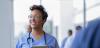 Female nurse or doctor smiles while staring out window in hospital hallway and holding digital tablet with electronic patient file