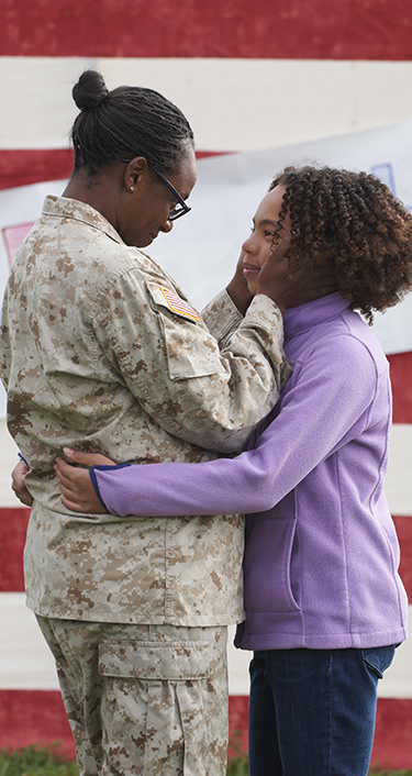 mother in military uniform hugging daughter with american flag in the background